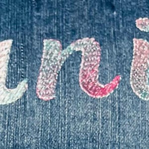 May include: A dark blue denim fabric featuring the name Danica embroidered in a cursive script font. The embroidery thread displays a gradient of colors, transitioning between shades of pink, light blue, and white across the letters.