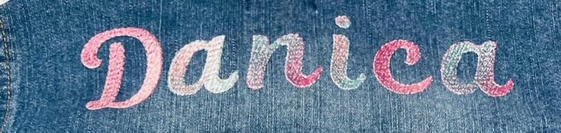 May include: A dark blue denim fabric featuring the name Danica embroidered in a cursive script font. The embroidery thread displays a gradient of colors, transitioning between shades of pink, light blue, and white across the letters.