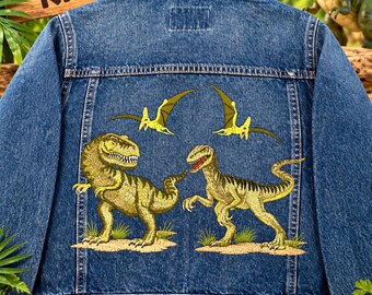 May include: A blue denim jacket featuring an embroidered back panel with two green dinosaurs and two flying pterosaurs. The jacket has a classic collar, long sleeves, and visible stitching. Above the jacket, a wooden sign reads DINO KID.