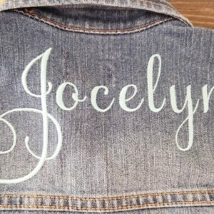 May include: A blue denim jacket featuring the name Jocelyn embroidered in white cursive script across the back. The garment includes a classic collar and visible stitching details along the seams, showcasing a textured fabric finish.