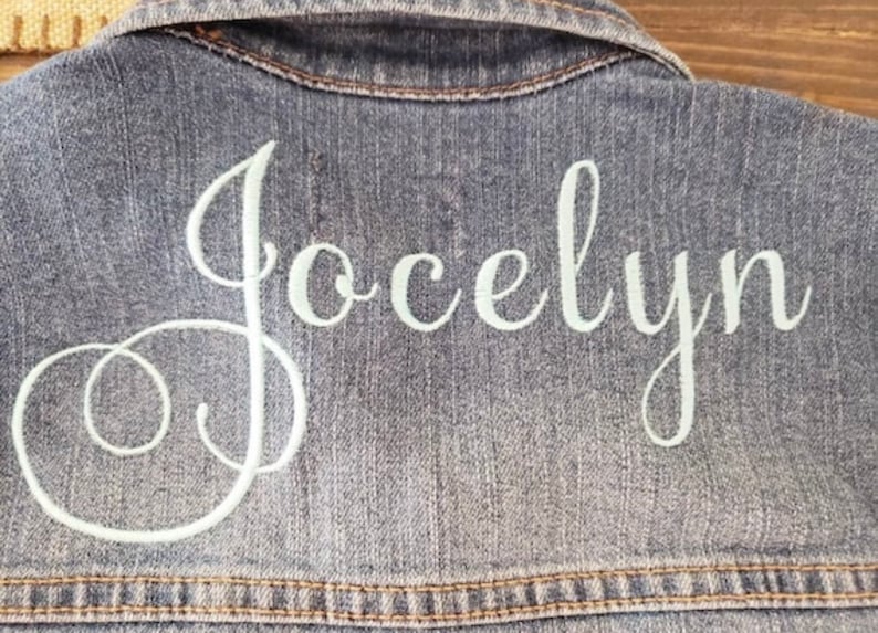 May include: A blue denim jacket featuring the name Jocelyn embroidered in white cursive script across the back. The garment includes a classic collar and visible stitching details along the seams, showcasing a textured fabric finish.