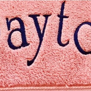 May include: A soft, light pink towel featuring the name Payton embroidered in dark navy blue thread. The text is centered and written in a clear, serif font, showcasing the textured fabric of the towel and the stitching detail.