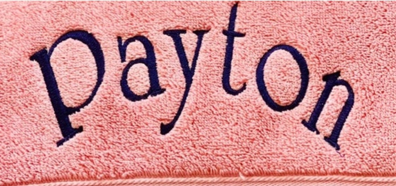 May include: A soft, light pink towel featuring the name Payton embroidered in dark navy blue thread. The text is centered and written in a clear, serif font, showcasing the textured fabric of the towel and the stitching detail.