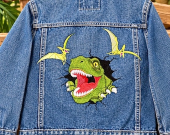 May include: A blue denim jacket featuring a large embroidered graphic on the back. The design shows a green T-Rex head emerging from a black crack, flanked by two yellow flying pterodactyls. The jacket has a classic collar and metal buttons.
