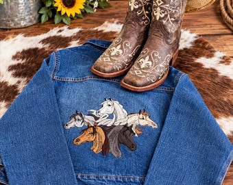 May include: A blue denim jacket featuring an embroidered design of five horse heads in various colors on the back. A pair of brown leather cowboy boots with white floral embroidery and a straw cowboy hat are positioned on top of the jacket.