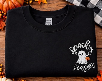 May include: A black crewneck sweatshirt featuring an embroidered white ghost holding a small orange jack-o'-lantern. The text "spooky season" is embroidered in white cursive script above and below the ghost. A Gildan brand label is visible.