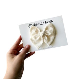 Könnte beinhalten: Eine cremefarbene Schleife mit gestickten floralen Details, präsentiert auf einer weißen Karte. Die Karte zeigt den Text "all the cute bows" in handschriftlichem Stil. Die Website-Adresse @thecutebowsettsy.com ist ebenfalls sichtbar.