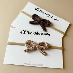 Puede incluir: Dos diademas para bebés con lazos de ganchillo. Un lazo es marrón oscuro, el otro es marrón claro. Cada lazo está unido a una banda elástica fina y se muestra en una tarjeta blanca con el texto "all the cute bows". La dirección del sitio web allthecutebowsetsy.com está impresa.
