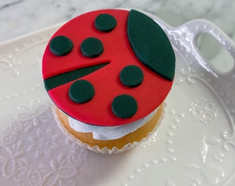 May include: A red and green ladybug cupcake with white frosting. The cupcake is on a white plate with a decorative pattern.