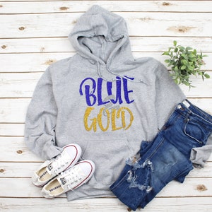 May include: A gray hoodie with the words "BLUE GOLD" in blue and gold glitter. The hoodie is paired with blue jeans and white sneakers. The items are arranged on a light wood-grain surface.