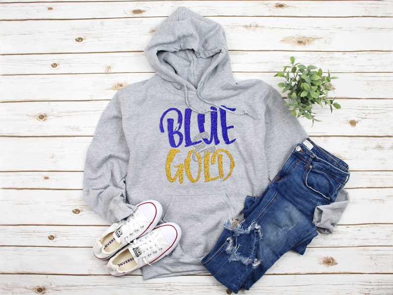 May include: A gray hoodie with the words "BLUE GOLD" in blue and gold glitter. The hoodie is paired with blue jeans and white sneakers. The items are arranged on a light wood-grain surface.