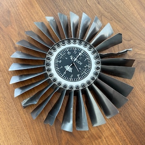 May include: A circular wall clock featuring a black face designed to resemble an aircraft altimeter with the word ALTITUDE printed in white. The clock is surrounded by a ring of metallic, dark gray turbine blades secured with bolts.