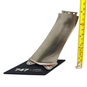May include: A silver aircraft blade, approximately 30 cm long, mounted on a black base with the text "747" and the Boeing logo. A yellow ruler is positioned next to the blade for scale.