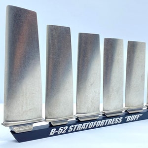May include: Six metal blades, possibly turbine blades, mounted on a black base. The base is labelled "B-52 STRATOFORTRESS 'BUFF'". A ruler to the left shows measurements in inches, with the blades appearing to be approximately 12 cm tall.