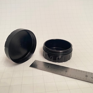 May include: A black metal container with a lid. The container is about 2 inches in diameter and 1 inch tall. The lid has a slightly raised edge.
