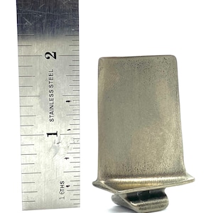 May include: A silver-colored metal turbine blade, approximately 2 inches in length, is shown next to a ruler.
