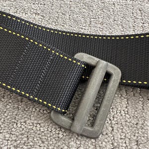 Puede incluir: Primer plano de una correa de nailon negro con costuras amarillas y una hebilla de metal gris. La correa es gruesa y duradera, diseñada para uso intensivo. La hebilla es rectangular y parece resistente.