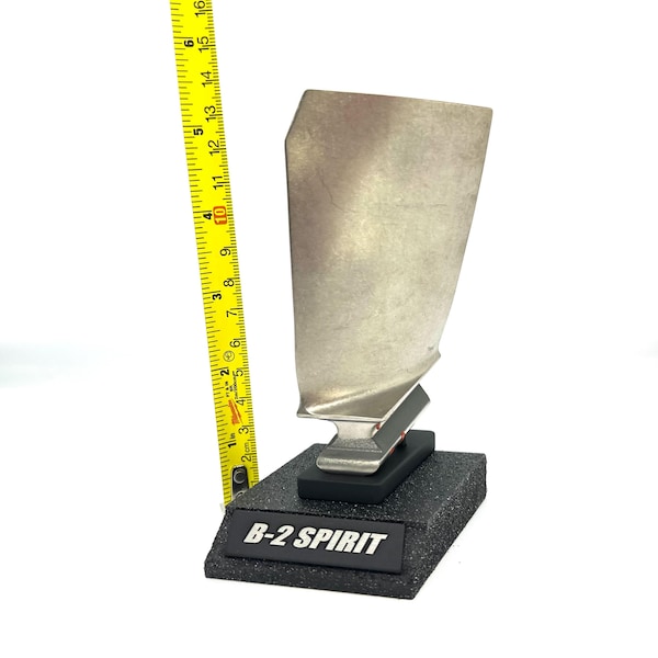 May include: A silver-colored metal turbine blade mounted on a black base with the text "B-2 SPIRIT". The blade is approximately 10 inches tall, as measured by a yellow tape measure.