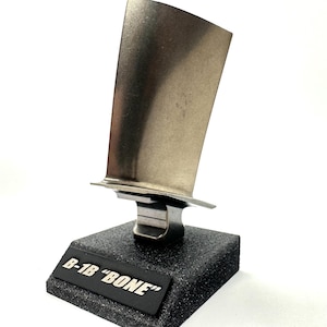May include: A silver metal model of a jet engine blade on a black base with the text "B-1B "BONE" printed on a silver plaque.
