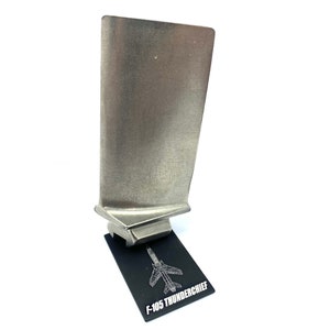 May include: A silver metal model of a jet engine on a black stand with the text "F-105 THUNDERCHIEF" printed on it.
