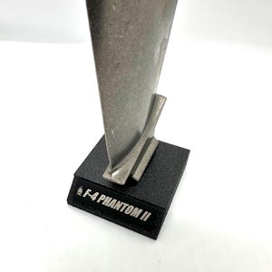 May include: A silver metal model of an F-4 Phantom II jet engine on a black base. The base has the words "F-4 PHANTOM II" printed on it.