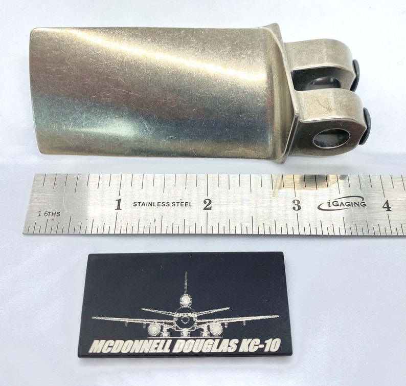 May include: A metallic silver-colored aircraft part with a ruler showing inches. A black rectangular plaque with a white illustration of an airplane and the text "MCDONNELL DOUGLAS KC-10" is below the ruler.
