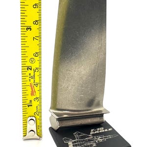 May include: A metallic turbine blade, approximately 9 inches tall, is displayed on a black base with an F-15 Eagle graphic. A yellow tape measure is positioned next to the blade, showing inches and centimeters.