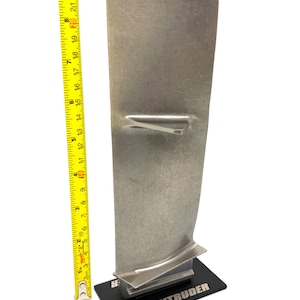 Puede incluir: Un álabe de turbina de motor a reacción A-6 Intruder metálico montado sobre una base rectangular negra. Una cinta métrica amarilla colocada verticalmente junto al álabe indica una altura de aproximadamente 25 cm desde la base hasta el borde superior.