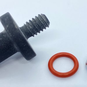 Puede incluir: Un tornillo de metal negro con un bucle en el lateral, una junta tórica de goma roja y una tuerca de metal plateado con una arandela de goma morada.