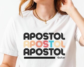 May include: White t-shirt with the word "APOSTOL" in a stacked design, with each word in a different color. The word "Guitar" is below the design. The person is wearing blue jeans.