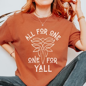 May include: A rust-colored t-shirt with the text "ALL FOR ONE ONE FOR Y'ALL" in white. The shirt features a stylized design above the words. The person is wearing blue jeans and white sneakers.