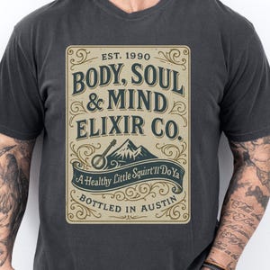 May include: Dark gray t-shirt featuring a vintage-style graphic. The graphic reads "BODY, SOUL & MIND ELIXIR CO." with additional text: "A Healthy Little Squirt'll Do Ya" and "BOTTLED IN AUSTIN". The design includes a mountain and musical instrument.
