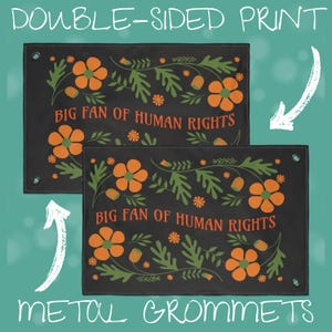 Close-up graphic showing the “Big Fan of Human Rights” house flag with a double-sided print. Two views of the same flag are shown to demonstrate that the floral folk design and text appear clearly on both sides. The flag features a black background with orange flowers and green leafy motifs, reinforced stitching, and metal grommets in the corners for secure hanging on a flagpole or outdoor display.