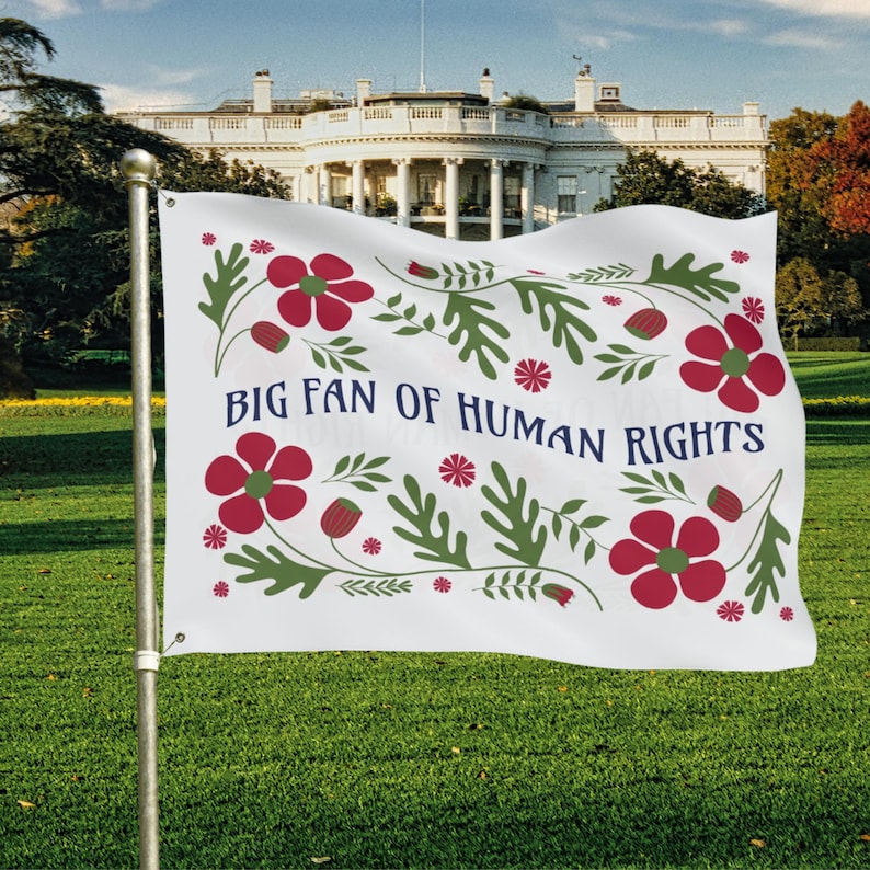 White full-size house flag displayed on a porch-style flagpole, featuring the phrase “Big Fan of Human Rights” in navy blue lettering. The design is surrounded by folk-inspired red flowers and green leafy motifs in a red, white, and blue color palette. Lightweight double-sided fabric waves in the breeze, creating a bold yet welcoming social justice and equality statement suitable for front-of-home or outdoor display.