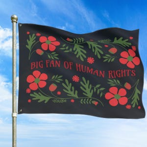 Black full-size house flag shown flying on a porch-style flagpole against a blue sky, featuring the phrase “Big Fan of Human Rights” in red lettering. Folk-inspired red flowers and deep green leafy motifs frame the message, creating a bold, high-contrast design. Lightweight double-sided fabric waves in the breeze, making this a striking social justice, equality, and peaceful protest flag for outdoor home display.