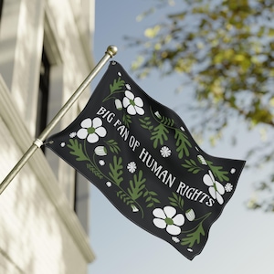 May include: A black flag with the text "BIG FAN OF HUMAN RIGHTS" in white. The flag is decorated with white flowers, green leaves, and small white star-shaped accents. The flag is attached to a silver pole.