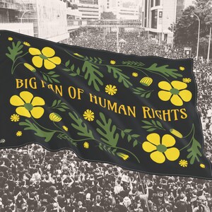 Black “Big Fan of Human Rights” house flag shown waving above a large crowd at a peaceful protest. The flag features bold yellow flowers and green leafy folk motifs framing the text, creating strong contrast against the dark background. Lightweight fabric moves in the breeze, presenting the design as a visible social justice, equality, and protest statement suited for public demonstrations or outdoor display.