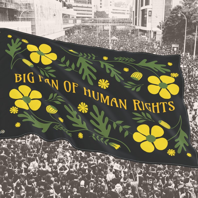 Black “Big Fan of Human Rights” house flag shown waving above a large crowd at a peaceful protest. The flag features bold yellow flowers and green leafy folk motifs framing the text, creating strong contrast against the dark background. Lightweight fabric moves in the breeze, presenting the design as a visible social justice, equality, and protest statement suited for public demonstrations or outdoor display.