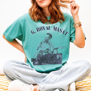 May include: Teal-colored t-shirt with the text "G. ROYAL MASAT" above a black and white graphic of a person playing a guitar. The shirt is a relaxed fit.