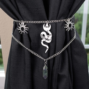 May include: A silver-colored curtain tieback featuring a chain design with sun and snake charms. A crystal pendant hangs from the bottom chain. The tieback is draped over a black curtain.