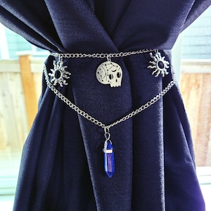 May include: A silver-colored curtain tieback featuring a chain design with sun and skull charms. A blue crystal pendant hangs from the chain. The tieback is draped over a dark blue curtain, creating a decorative accent.