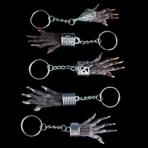 Puede incluir: Cinco llaveros con pequeñas manos oscuras con uñas largas y puntiagudas. Cada mano está unida a un anillo plateado mediante una cadena y un brazalete decorativo. Los brazaletes tienen varios diseños, incluyendo filigranas y patrones geométricos.