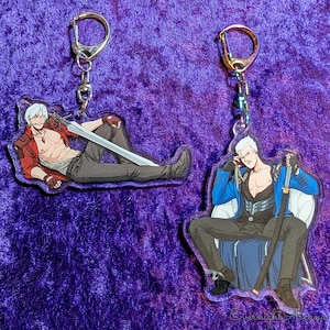 Op de afbeelding: Twee acryl sleutelhangers met illustraties van anime-personages. Eén sleutelhanger toont een personage in een rode jas, met een zwaard. De andere toont een personage in een blauwe jas, zittend met een zwaard. Beide sleutelhangers hebben zilverkleurige hardware.