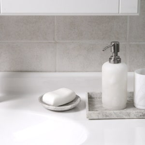 May include: A white ceramic soap dispenser with a silver pump, a white bar of soap in a gray ceramic dish, a white ceramic toothbrush holder with a white toothbrush, and a gray marble tray.