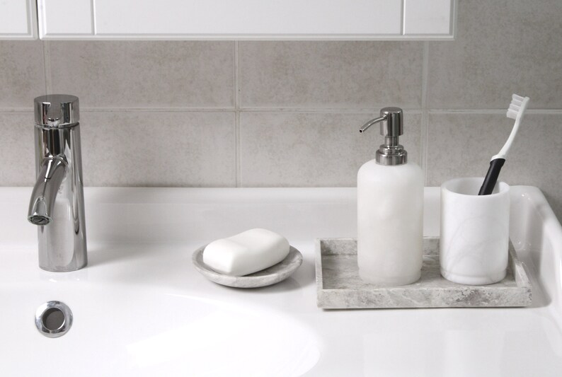 May include: A white ceramic soap dispenser with a silver pump, a white bar of soap in a gray ceramic dish, a white ceramic toothbrush holder with a white toothbrush, and a gray marble tray.