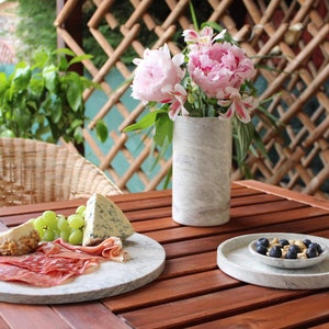 May include: A wooden table with a marble serving tray filled with cheese, grapes, and prosciutto. A smaller marble serving tray with blueberries and nuts sits on the table. A vase with pink flowers sits on the table.