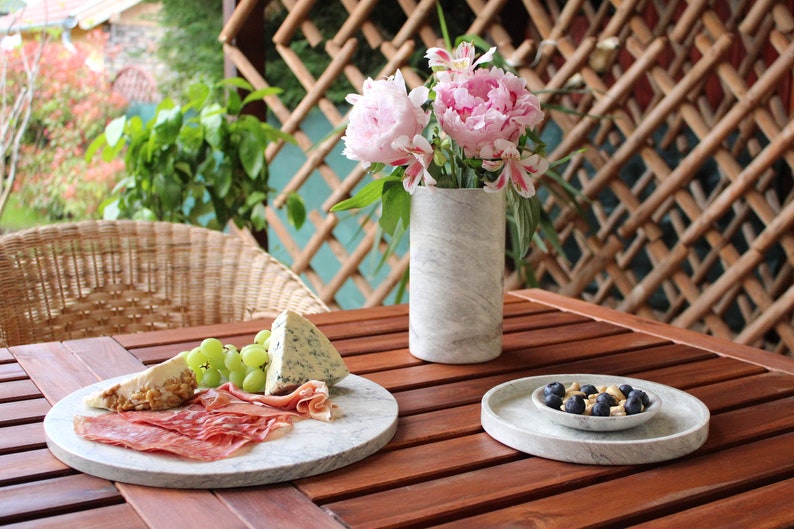 May include: A wooden table with a marble serving tray filled with cheese, grapes, and prosciutto. A smaller marble serving tray with blueberries and nuts sits on the table. A vase with pink flowers sits on the table.