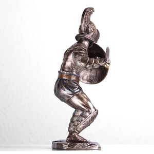 May include: A bronze statue of a gladiator in a fighting pose, holding a sword and shield. The statue is detailed with gold accents and is standing on a rocky base.