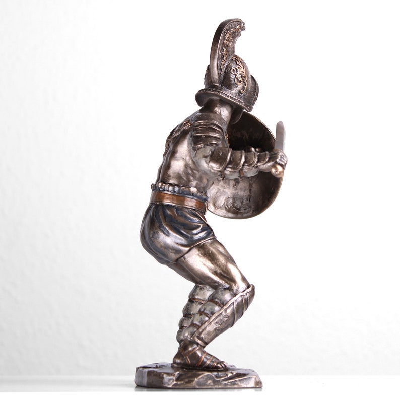 May include: A bronze statue of a gladiator in a fighting pose, holding a sword and shield. The statue is detailed with gold accents and is standing on a rocky base.