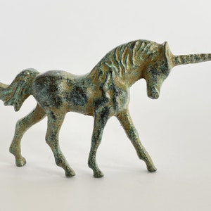 May include: A small, green and gold metal unicorn figurine. The unicorn is standing on all four legs with its head turned to the side. The unicorn has a long, thin horn.